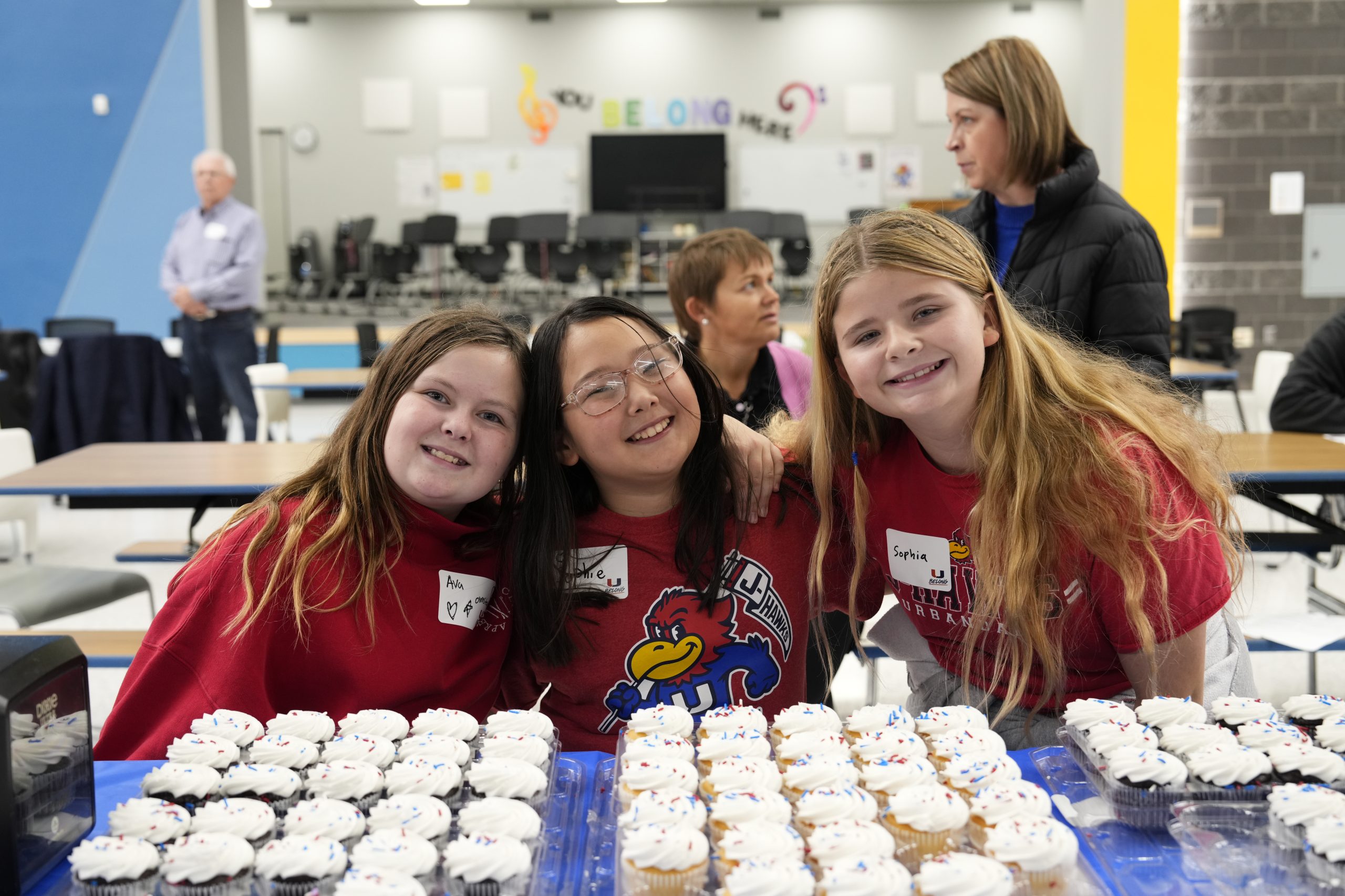 Three young students smiling in cafeteria behind a table of cupcakes