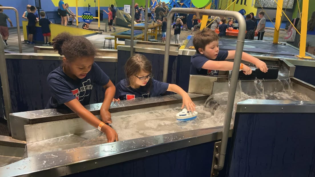 Three elementary students play with a river simulation at the science center. The student in the middle places a toy boat in the water and the two other students create dams with black plastic slabs.