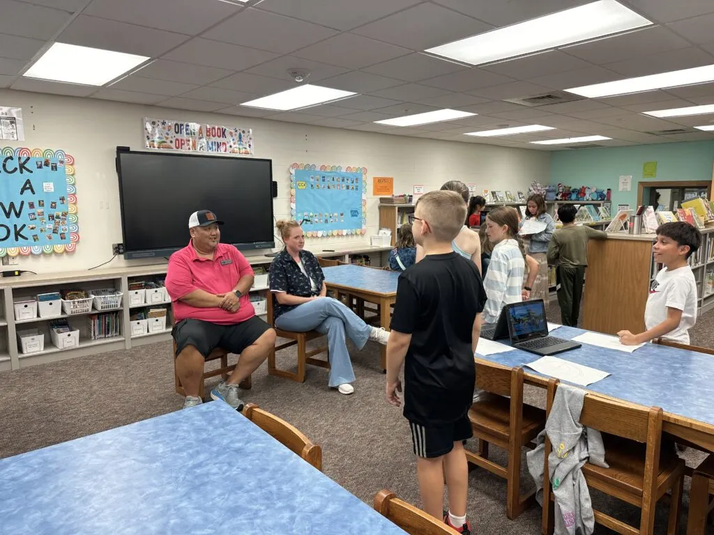 Two adults sit and listen to a small group of students present a class project.