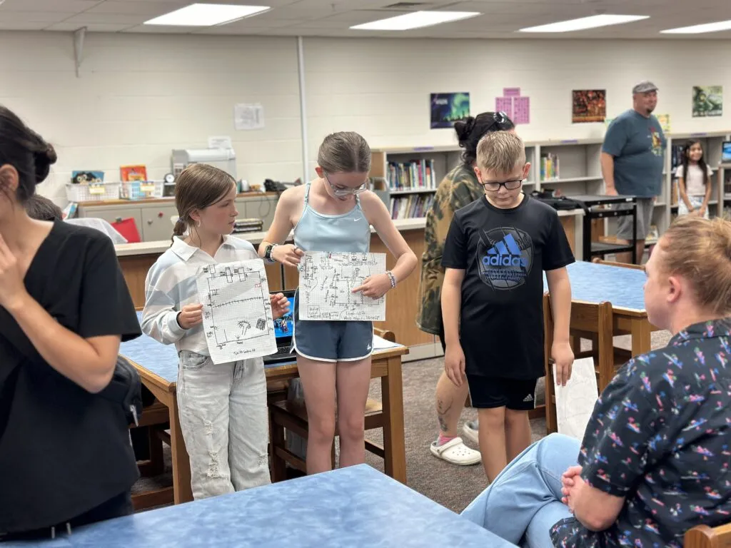 Three students present a class project. They hold hand drawn diagrams on graph paper.