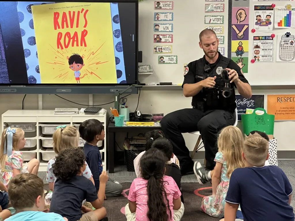 Police officer sits in front of an elementary school class. To his right is a TV screen showing the cover for picture book 