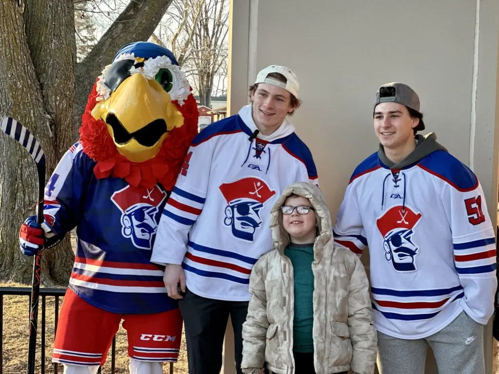 Des Moines Buccaneers mascot and hockey players in jerseys stand and smile with an elementary student