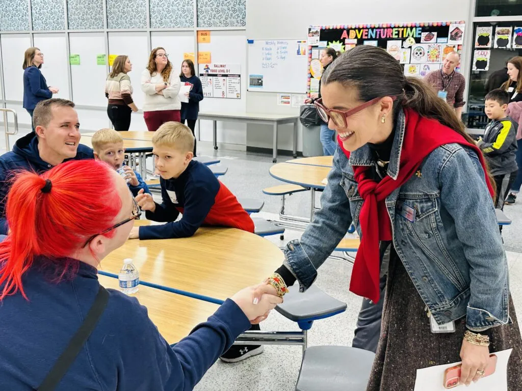 A teacher shakes hands with a student's family member in a school cafeteria.