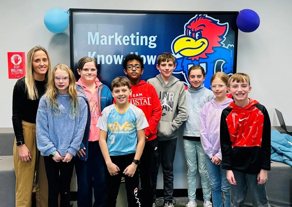 Eight elementary students and an adult stand and smile in front of a TV screen that says 