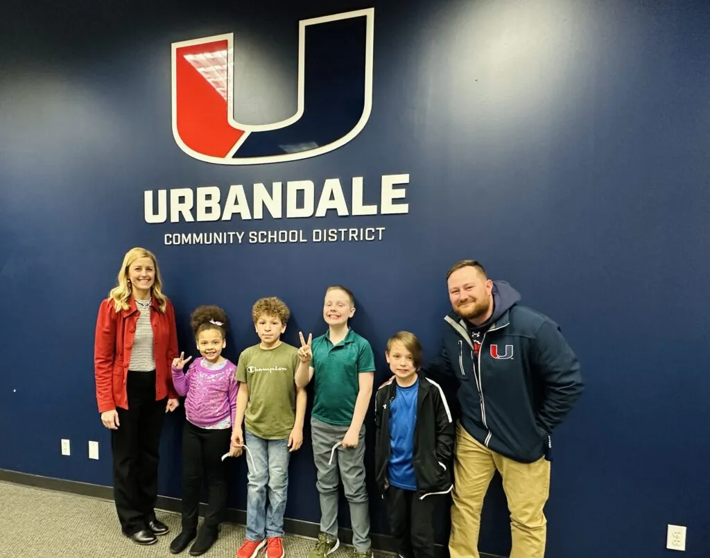 Four elementary students and two adults stand and smile in front of a blue wall with the UCSD logo. Two students hold up peace signs with their hands.