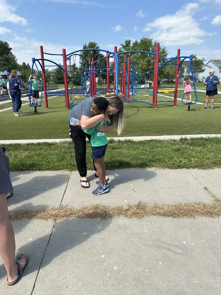 A staff member hugs an elementary student. There is a red and blue playground set in the background where other children play.