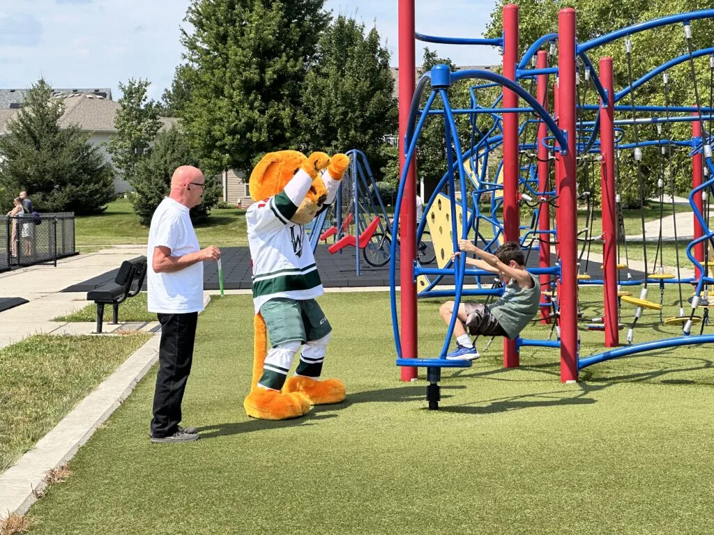 Iowa Wild mascot Crash, a wildcat wearing a green and white hockey uniform, cheers for an elementary student playing on playground equipment. A staff member stands nearby.