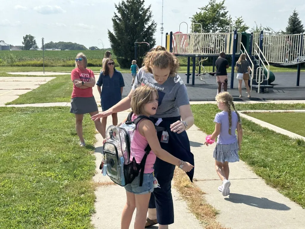 An elementary student approaches a teacher for a hug on the playground.