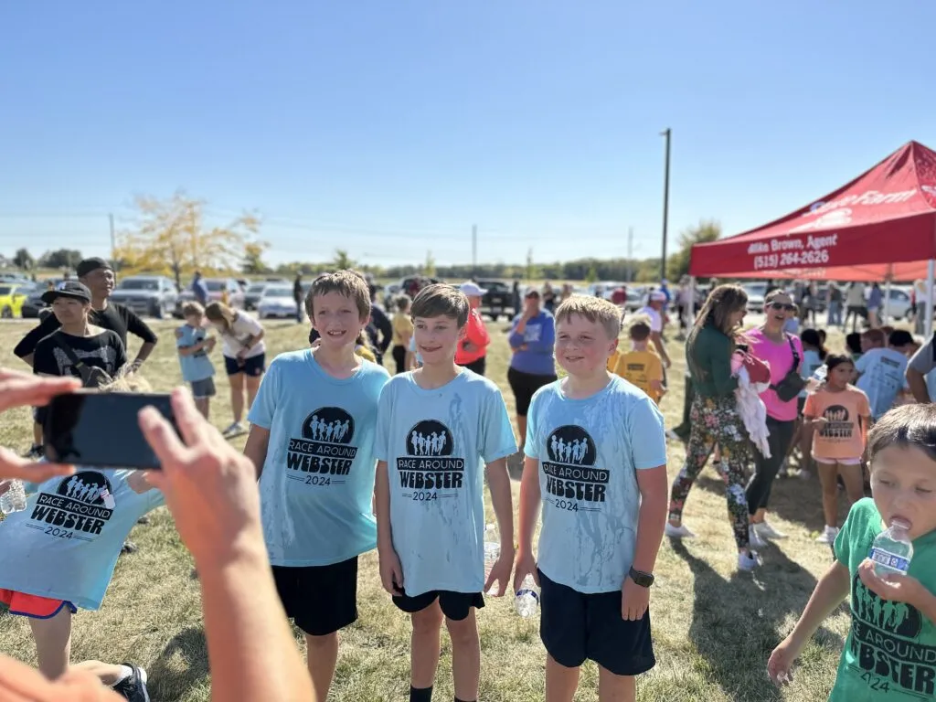 Three elementary students in light blue shirts stand together and smile while someone off camera takes a picture on their phone.