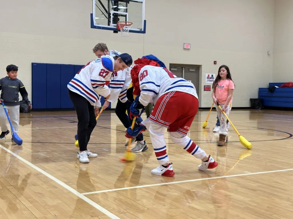 Des Moines Buccaneers hockey players and the Buccs mascot play hockey with elementary students in a school gym.
