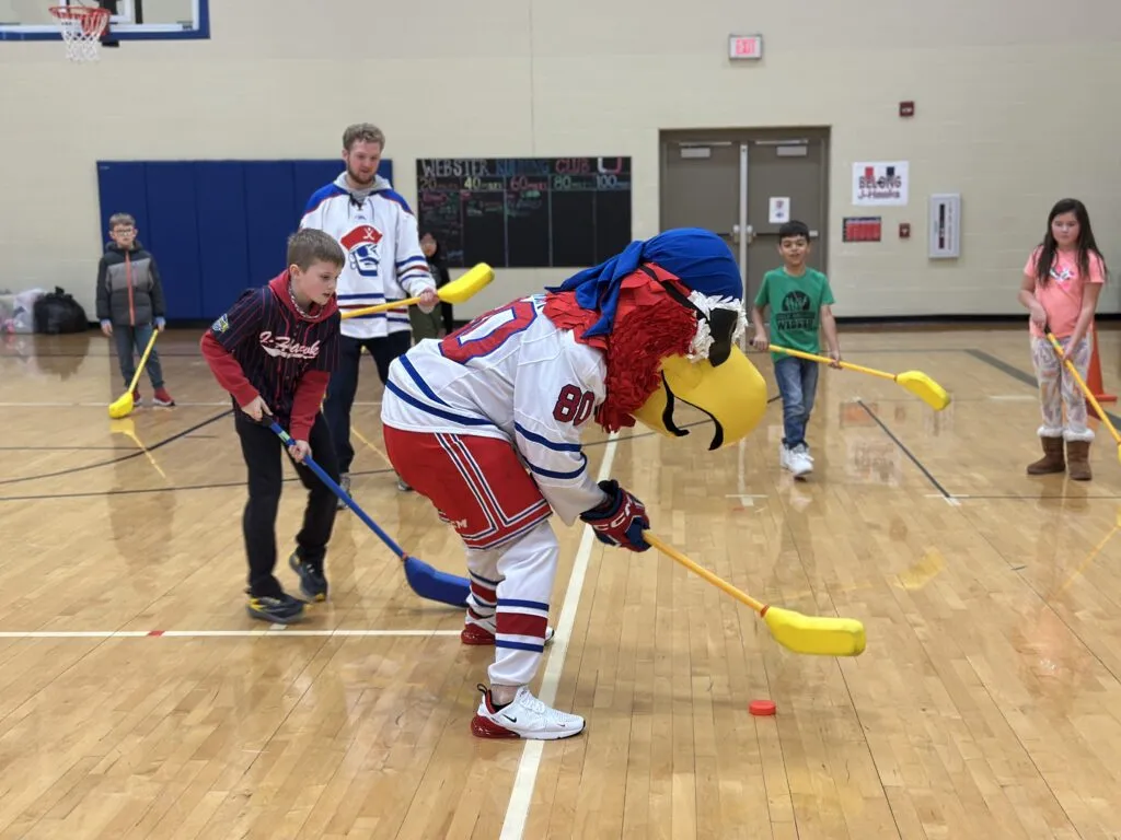 Des Moines Buccaneers hockey players and the Buccs mascot play hockey with elementary students in a school gym.