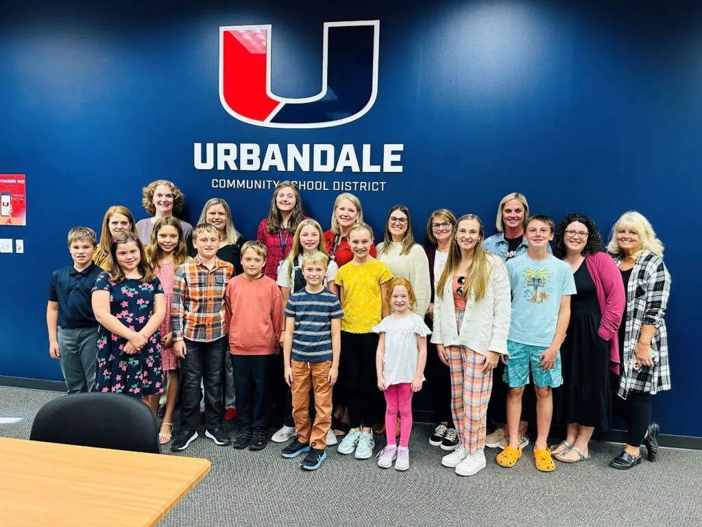 A group of Webster elementary students and adults stand and smile in front of a blue wall with the UCSD logo.