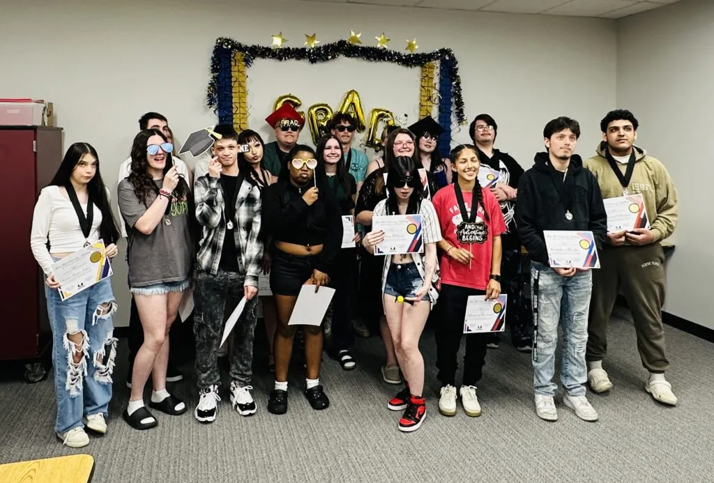 A large group of graduating students pose in front of a backdrop with yellow and blue streamers and balloons that spell 