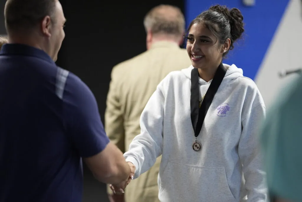 A student wearing a medal shakes hands with a staff member.
