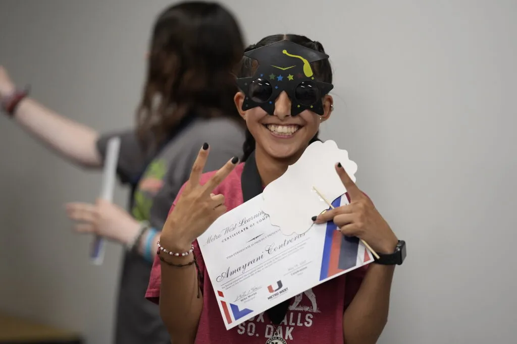 A student holds a Metro West Certificate of Completion and wears graduation-themed sunglasses. The student holds. up a peace sign.
